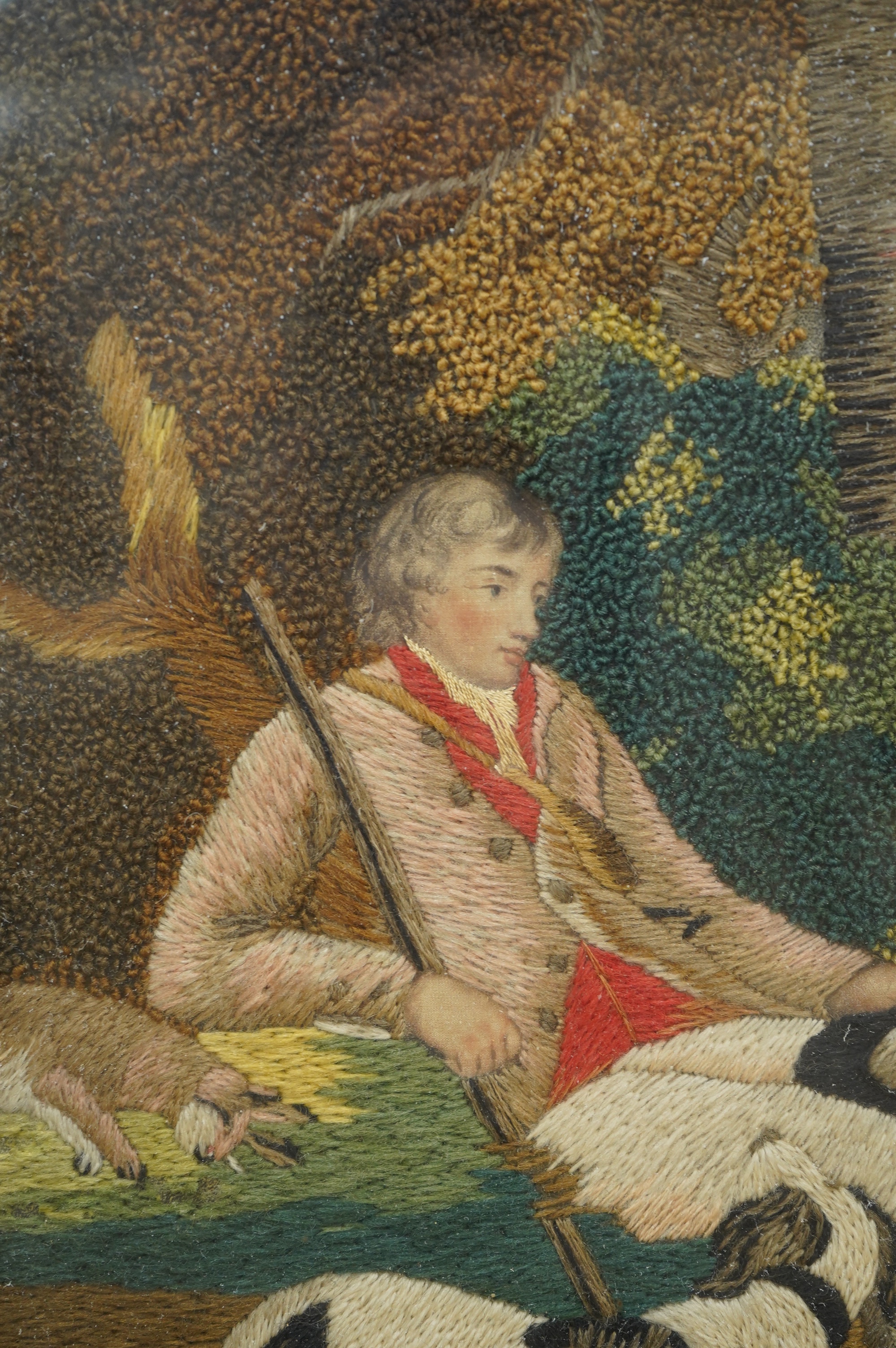 A framed early 19th century wool worked embroidery of huntsman sitting with a rabbit and his three hunting dogs under a tree, by a stream, with a field and cottage in the background, the embroidery worked in fine multi-c